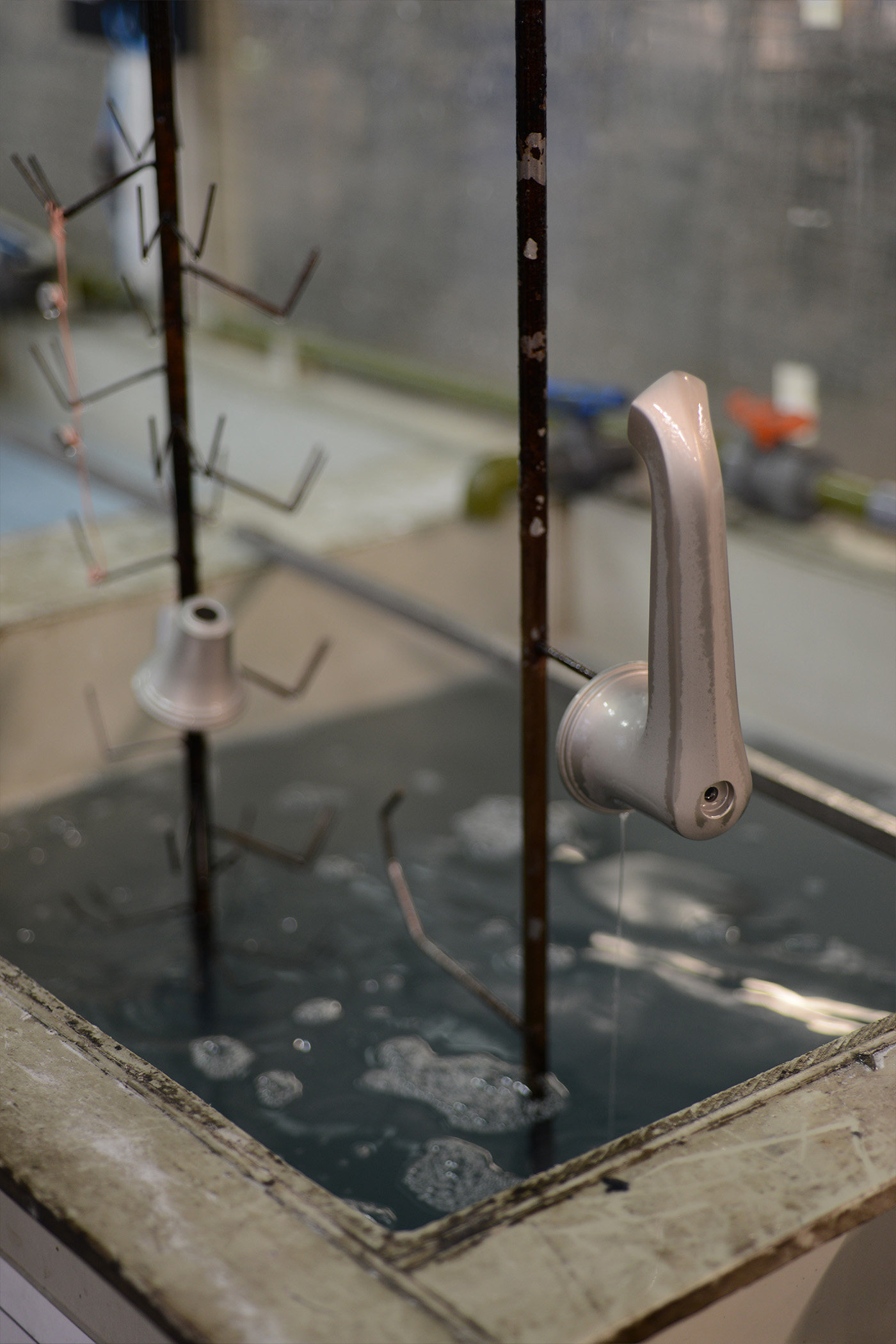 A basin spout and side valve parts on a jig being lifted from a cleaning solution bath to rinse excess lacquer