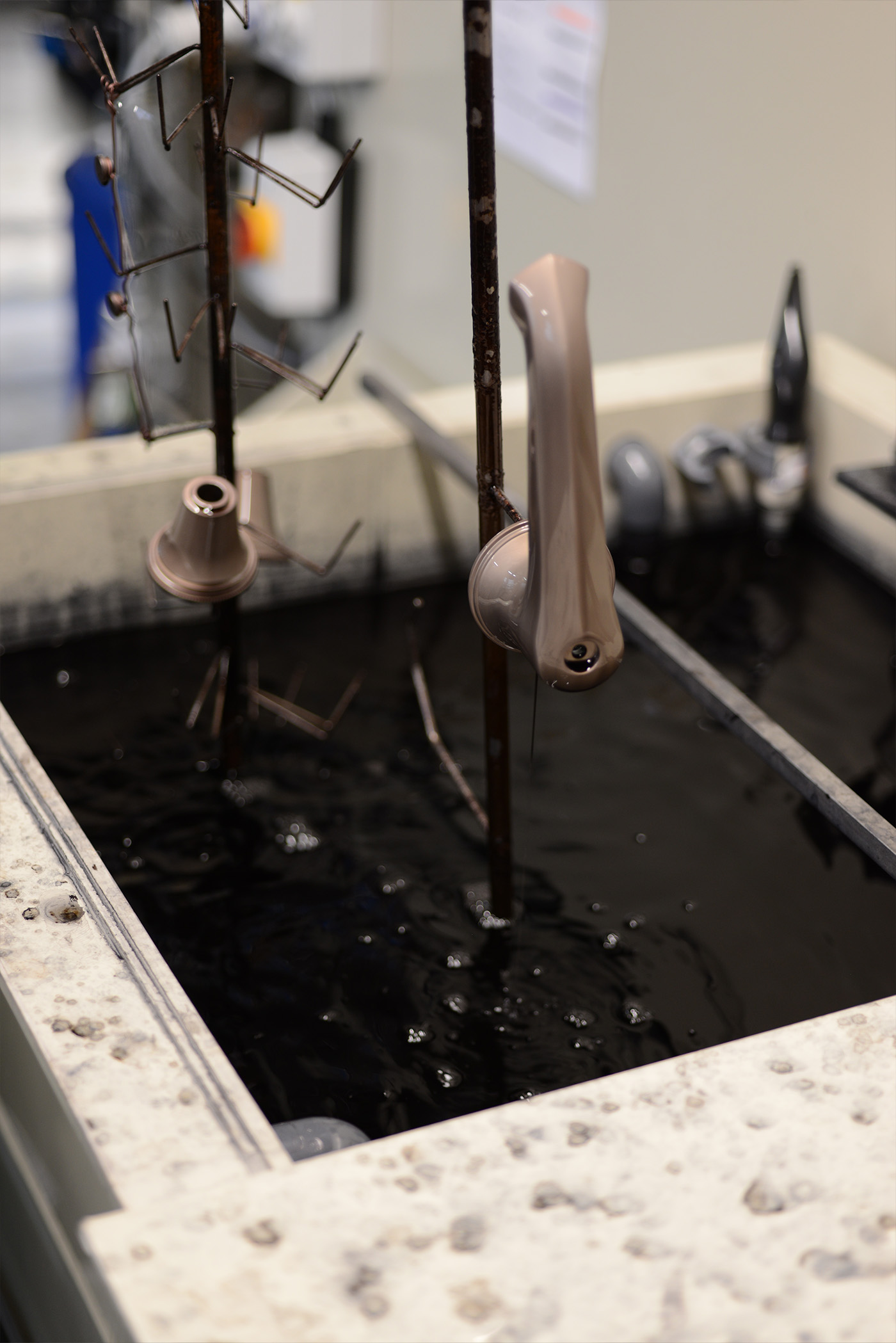 A basin spout and side valve parts on a jig being lifted from a dark dye bath which gives its multi-tonal appeal